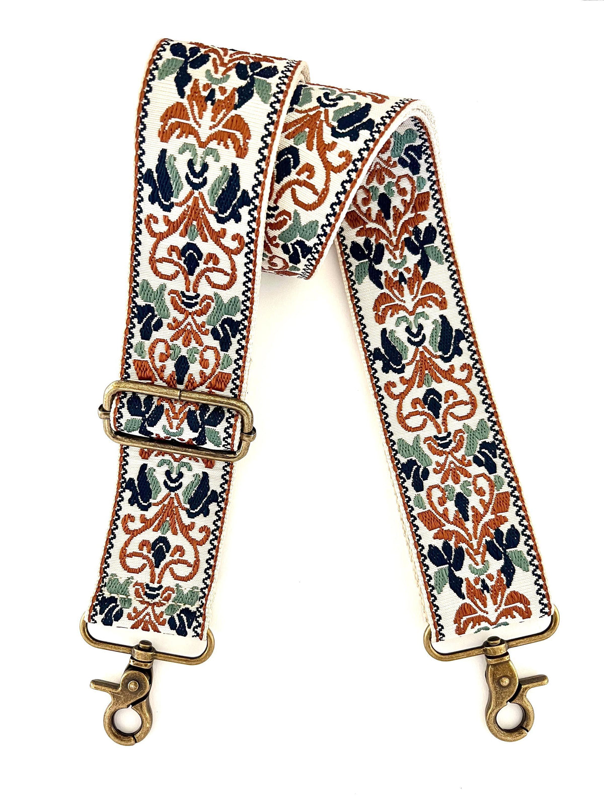 A wide fabric shoulder strap with an ornate floral and scroll pattern in rust orange, navy blue, sage green, and cream, finished with antique brass hardware clasp ends.
